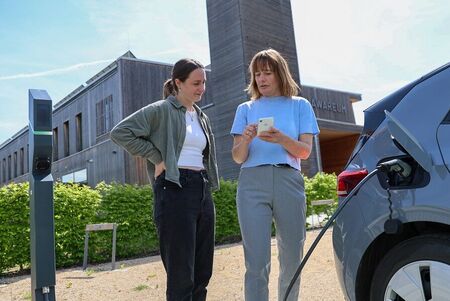 Zwei Frauen schauen auf ein Handy während des Ladens eines Elektroautos. Das Elektroauto steht rechts, die Ladesäule links und im Hintergrund befindet sich ein imposanten Holzgebäude mit Turm.