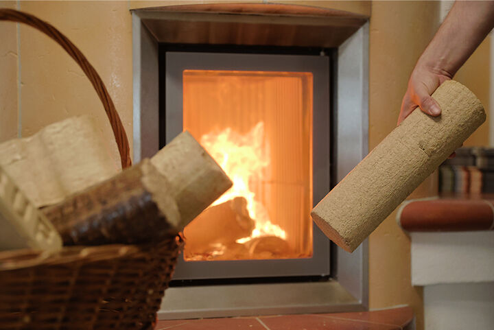 Hand hält ein Holzbrikett vor einem Ofen in dem ein Feuer brennt